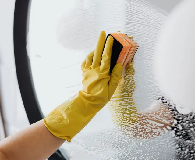 A yellow-gloved hand scrubbing glass with soapy sponge